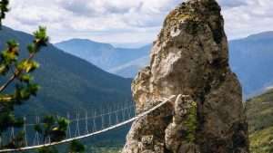 A rope bridge leads to a large rock formation with mountains in the background.