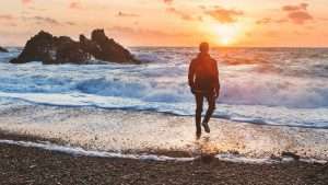 A person stands on the shore looking at the sunset.