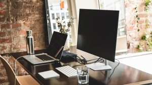 A black desk has a laptop, computer monitor, keyboard, and a glass of water on it with a brick wall and mirror in the background.