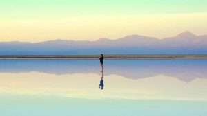 Someone stands on a thin body of water with their reflection and the reflection of the mountains below.