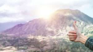 Someone holds their hand into the frame of the photo holding a thumbs up sign with the sun rising over a mountain in the background.