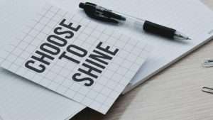 A piece of paper that reads, “Choose to Shine,” sits on top of a notebook and next to a pen.