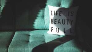 A green couch with a white pillow that has the phrase “Life is beauty full” on it.
