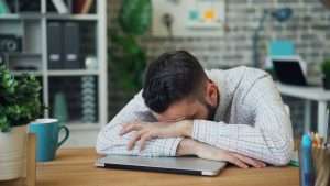A person resting their head on their arms folded over a laptop.