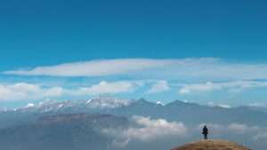 A person stands at the top of a mountain, looking out at even taller mountains in the distance.
