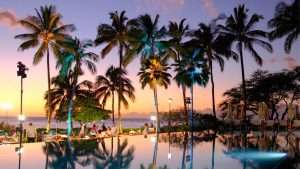 A pool surrounded by palm trees, near a beach, at sunset.